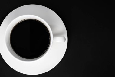 Siyah bir fonda, solda bir fincan çay tabağı üzerinde, siyah kahve dolu beyaz bir fincan duruyor.