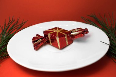 Kırmızı bir arka planda köknar dalları arasında beyaz bir fincan tabağı üzerinde şeker şeklinde bir Noel süslemesi bulunur.