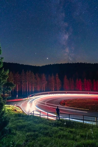 Milkyway, larga exposición de un hombre parado en una barandilla frente a una calle con curvas, mientras los coches pasan y dejan la luz del camino. — Foto de Stock