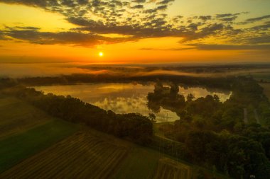 Gün doğumunun erken saatlerinde etrafı sisli ağaçlarla çevrili bir göl manzarası.