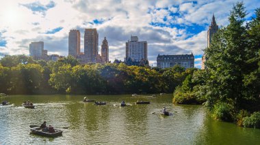 New York Central Park 'ta güzel bir gün, insanlar arka planda etkileyici gökdelenlerin önünde kayıkla geziyorlar..