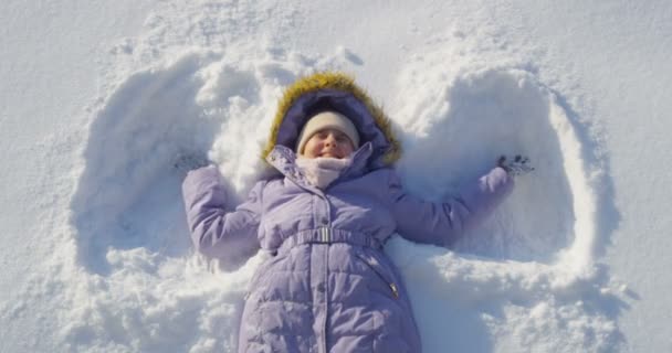 微笑的孩子躺在雪制造雪天使拍摄从顶部的角度在阳光一天高角度慢运动的手臂和腿躺在后面关闭 — 图库视频影像
