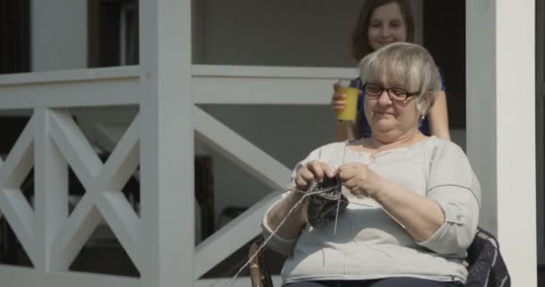 高齢の孫の世話は 赤いカメラで撮影されたテラッセのハウスポーチで晴れた日に一緒に編み物をして 一緒に笑顔をしている老祖母にオレンジジュースをもたらしています — ストック動画