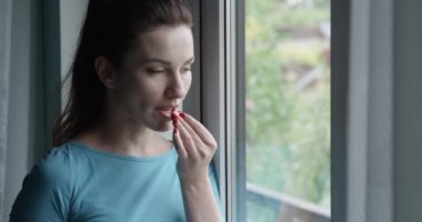 Bir bardak su içen ve hap alan kadın Pencere kenarında duruyor.