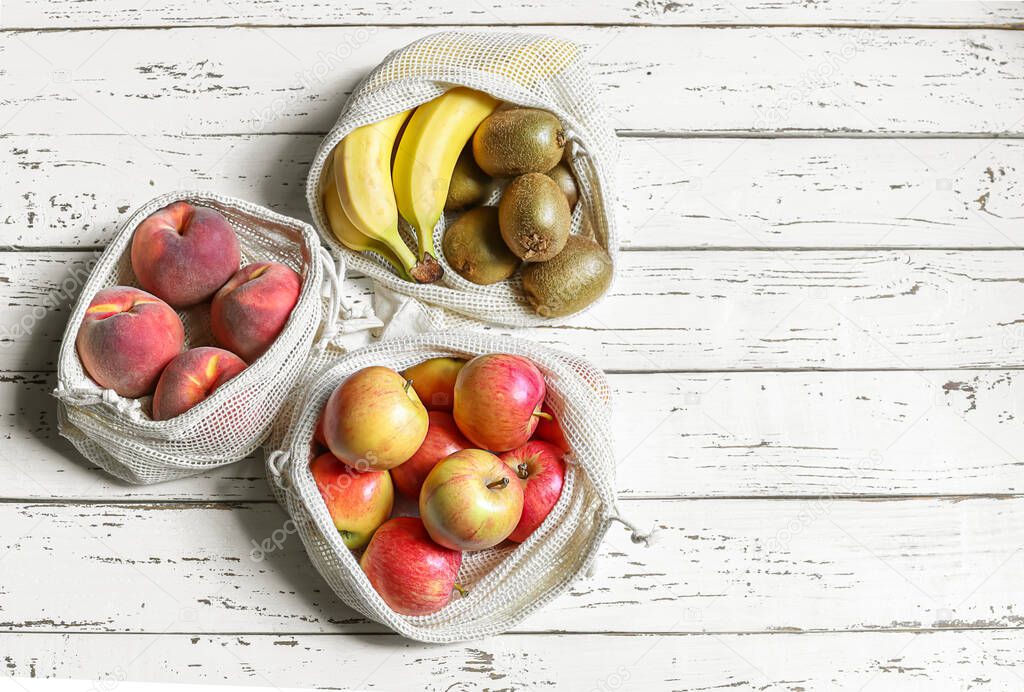 Apples, peach, kiwi, bananas in reusable eco friendly mesh textile bags on the white wooden background. Zero waste concept and plastic free lifestyle. Top view, copy space