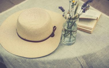 Yaban çiçekleri, açık bir kitap ve masa örtüsü, retro filtre, kulübe özünde estetik konsept ile kaplı bir masa üzerindeki ribonlu kadınlar için yaz şapkası.