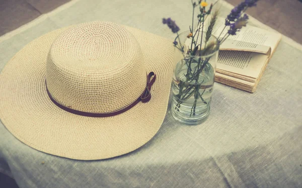 Yaban çiçekleri, açık bir kitap ve masa örtüsü, retro filtre, kulübe özünde estetik konsept ile kaplı bir masa üzerindeki ribonlu kadınlar için yaz şapkası.
