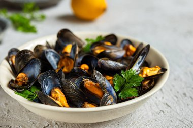 Beyaz bir tabakta beyaz şarap soslu enfes taze midyeler, gri bir arka planda bir limon ve maydanoz.