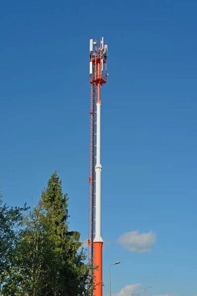 Tower with antennas of cellular communication. Against the blue sky and the top of a tree.