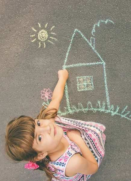 The child draws the house with chalk on the asphalt. Selective focus.