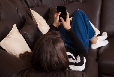 Oturma odasındaki kanepede rahat bir evde akıllı telefon kullanan genç bir kadın. Kanepede deri yastıklar var..