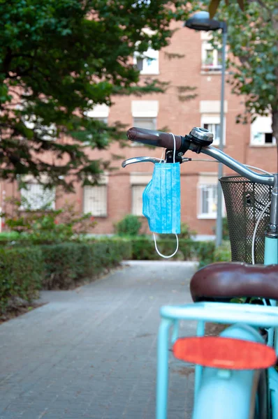 Surgical mask hanging from the handlebars of a bike parked outdoors and in the open air. Disposable masks without throwing away. Sky blue bike with mask