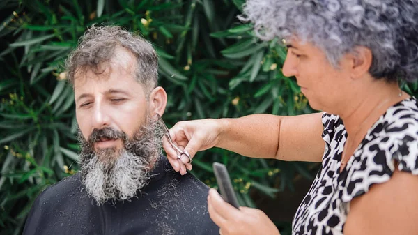Siyah ve beyaz sakallı beyaz bir adamın sakal tıraşı olurken çekilmiş resmi. Kuaför makas ve tarak kullanıyor..