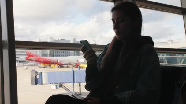 Giovane donna turistica di razza mista che utilizza il telefono cellulare vicino alla finestra al terminal dell'aeroporto di partenza. 4K . — Video Stock