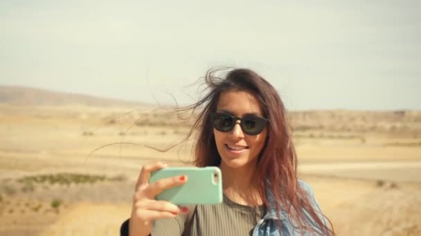 Молодий привабливою посміхається дівчина змішаної раси займає панорамну фотографію пустелі на мобільний телефон. Щасливі туристичних жінка в Sunglases запису відео. Каппадокії. 4 к Slowmotion. — стокове відео