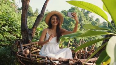 Genç mutlu Selfie fotoğrafları cep telefonuyla Dekoratif saman yuvada oturan yapma beyaz elbiseli yarış turist kız karışık. Yaşam tarzı seyahat 4k görüntüleri. Bali, Endonezya.