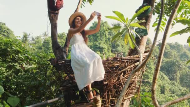 Junges fröhliches gemischtes Touristenmädchen in weißem Kleid, das Selfie-Fotos mit dem Handy macht und im dekorativen Strohnest sitzt. Lebensstil Reise 4k Filmmaterial. bali, indonesien. — Stockvideo