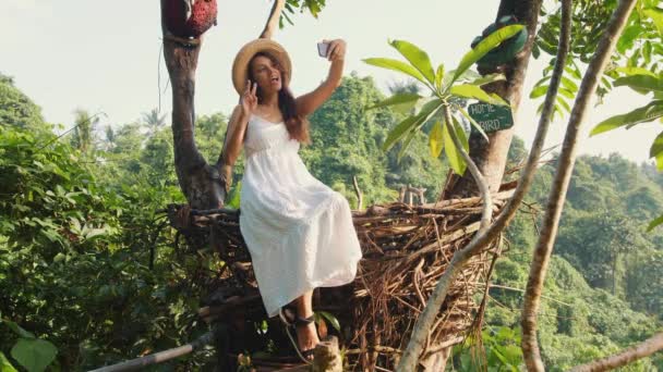 年轻快乐的混血旅游女孩在白色礼服制作自拍照片使用手机坐在装饰秸秆巢。生活方式旅游4k 视频。巴厘岛, 印度尼西亚. — 图库视频影像