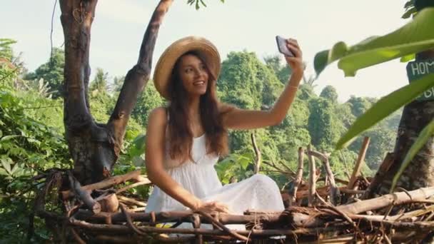 Young Smiling Mixed Race Tourist Girl in White Dress Making Selfie Photos Using Mobile Phone Sitting in Decorative Straw Nest. Lifestyle Travel 4K Footage. Bali, Indonesia. — Stock Video