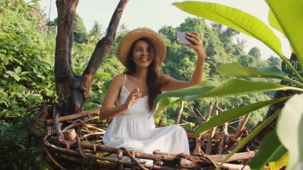年轻快乐的混血旅游女孩在白色礼服制作自拍照片使用手机坐在装饰秸秆巢。生活方式旅游4k 视频。巴厘岛, 印度尼西亚. — 图库视频影像