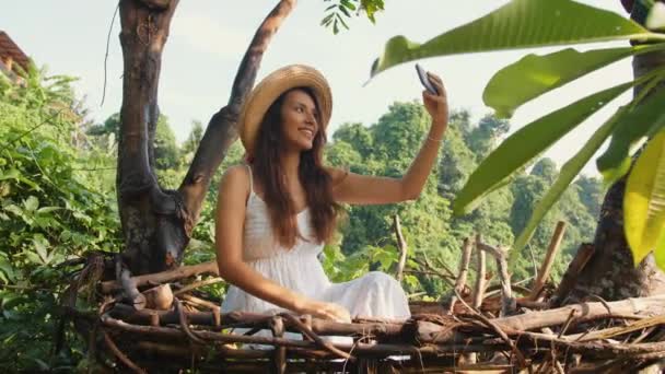 Молодая улыбающаяся смешанная раса туристка в белом платье делает селфи фотографии с помощью мобильного телефона, сидя в декоративной Соломенное гнездо. "Путешествие в стиле жизни" 4К. Бали, Индонезия . — стоковое видео