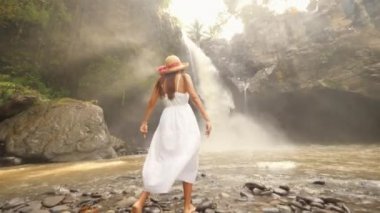 Genç kız beyaz elbise ve hasır şapka Tegenungan şelaleye yürüyüş ve silah kadar yükselterek. Kaygısız yaşam seyahat 4k Slowmotion görüntüleri. Bali, Endonezya.