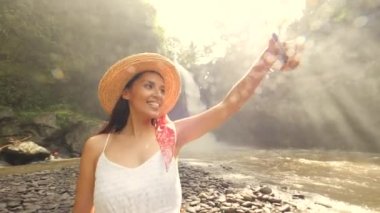 Gülümseyen genç yarış turist kız beyaz elbise ve hasır şapka Selfie fotoğrafları cep telefonuyla inanılmaz büyük Jungle Şelalesi ile yapma karışık. Yaşam tarzı seyahat 4k görüntüleri. Bali, Endonezya.
