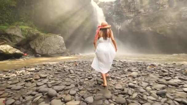 Młoda dziewczyna w białej sukni i słomy kapelusz Runing wodospad Tegenungan i podniesienie broni. Beztroski styl życia podróży 4k Slowmotion materiału. Bali, Indonezja. — Wideo stockowe
