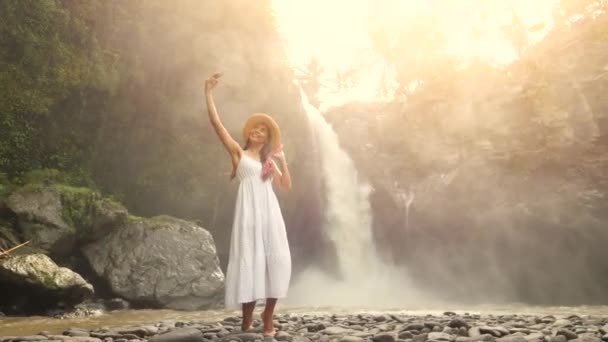 Joven sonriente mezcla de raza turista chica en vestido blanco y sombrero de paja haciendo fotos selfie usando el teléfono móvil con increíble cascada de la selva enorme. Estilo de vida Viajes 4K Filmación. Bali, Indonesia . — Vídeos de Stock