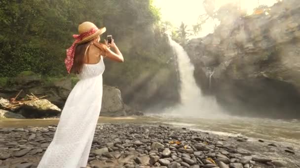 Hermosa joven turista en vestido blanco largo y sombrero de paja haciendo fotos de increíble cascada de Tegenungan usando teléfono inteligente móvil. Natural Lifestyle Travel 4K Slowmotion Footage (en inglés). Bali, Indonesia . — Vídeos de Stock