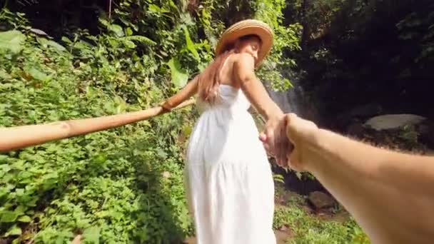 Joven atractiva chica de raza mixta en vestido blanco y sombrero de paja de la mano y el hombre principal a la increíble cascada de la selva. Estilo de vida Viaje 4K cámara lenta Gran angular POV Filmación. Bali, Indonesia . — Vídeos de Stock