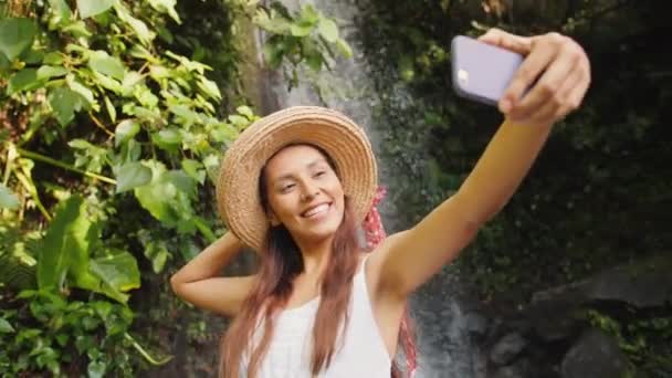 年轻快乐的混赛旅游女孩在白色礼服和草帽制作自拍照片使用手机与惊人的野生丛林瀑布。生活方式旅游4k 视频。巴厘岛, 印度尼西亚. — 图库视频影像