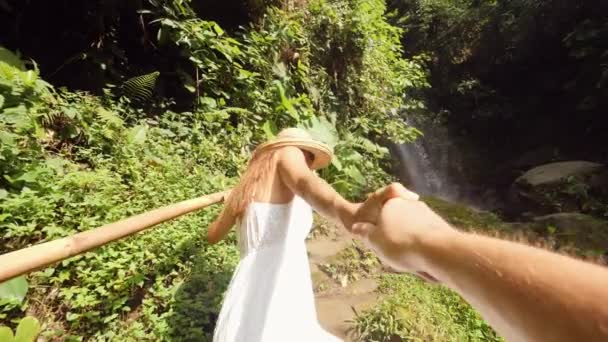 Junges lächelndes Mischlingsmädchen in weißem Kleid und Strohhut an der Hand und führt Mann zu erstaunlichem Dschungel-Wasserfall. Lifestyle Reisen 4k Zeitlupe Weitwinkel pov Filmmaterial. bali, indonesien. — Stockvideo