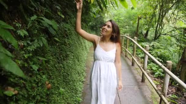 Jovem Menina Raça Mista Feliz em Vestido Branco Caminhando na Floresta da Selva, Olhando ao redor e tocando com as Folhas Tropicais Mão. Calm and Carefree Lifestyle Travel Filmagem em câmera lenta 4K. Bali, Indonésia . — Vídeo de Stock