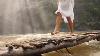 Çıplak ayakla küçük ahşap köprü üzerinde orman nehir tropikal yağmur ormanlarında yürüyüş beyaz elbiseli Genç kadın. 4k Slowmotion. Bali, Endonezya.