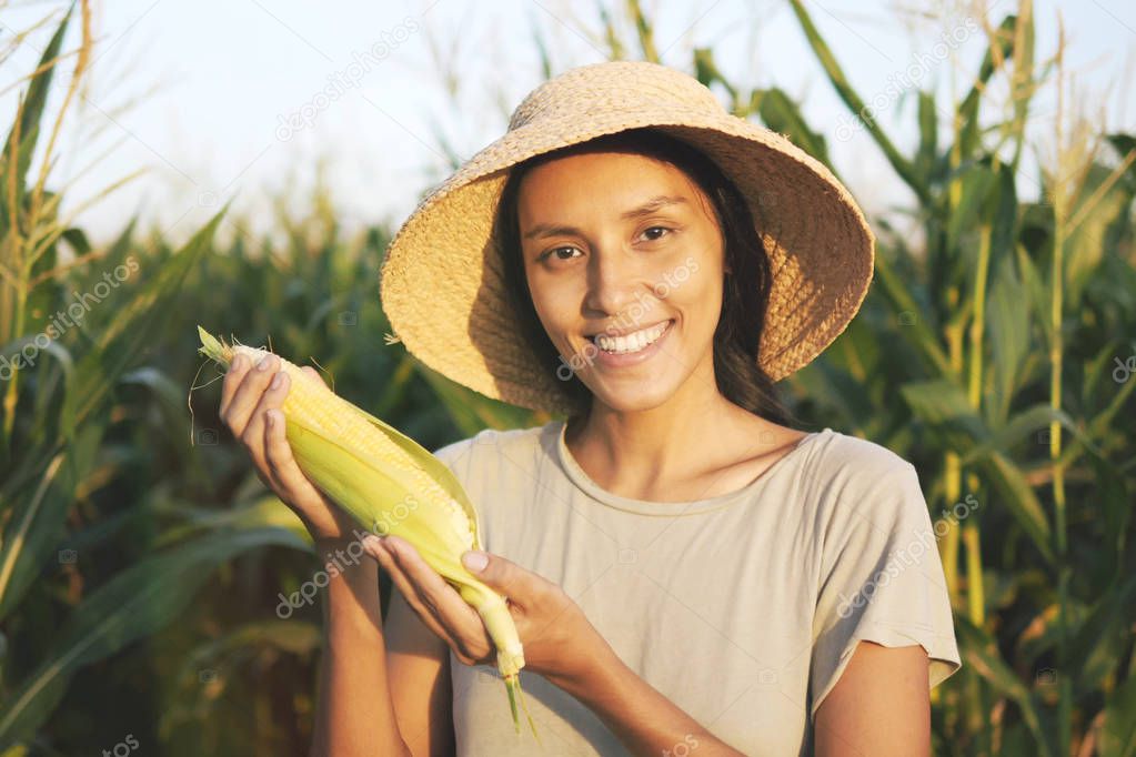 Beautiful Young Mixed Race Farmer Woman in Hat with Ripe Corn at Organic Farm Field. Woman Smiling and Showing Thumb Up at Camera in Natural Sunset Light. 4K, Slowmotion Agricultural Harvest Footage.