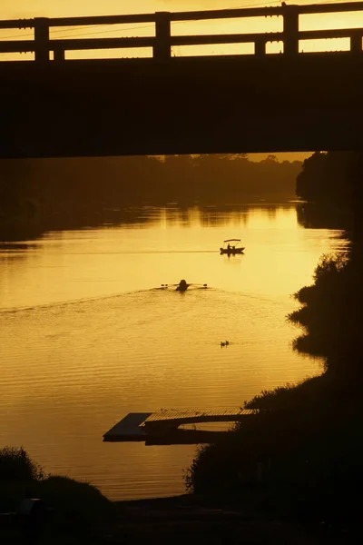 Rowers and safety boat silhouetted under a bridge in a golden sunrise on the river.