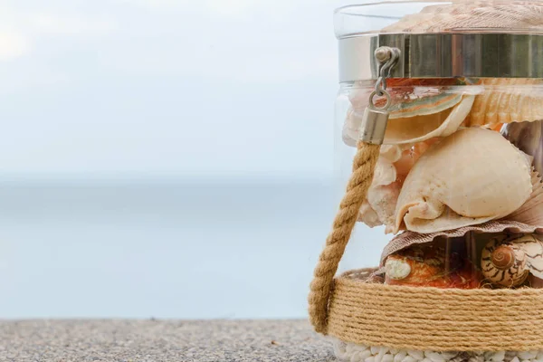Разнообразие морских раковин в стеклянной банке и морской пейзаж на заднем плане с копировальным пространством. Концепция отдыха — стоковое фото