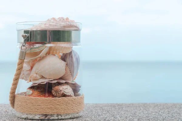 Разнообразие морских раковин в стеклянной банке и морской пейзаж на заднем плане с копировальным пространством. Концепция отдыха — стоковое фото