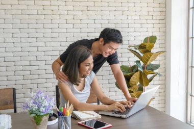 Genç Asyalı çift, modern mutfaktaki büyük ahşap masada otururken ve evde bilgisayar laptopuyla çalışırken rahat kıyafetler giyerler. Aşk, mutluluk, ev konseptinde çalışmak