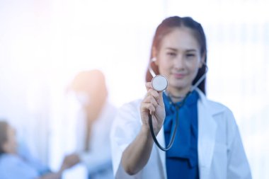 Stetoskop başı tutan genç bir kadın doktor ve arka plan üzerinde çalışan bulanık bir tıbbi ekip.