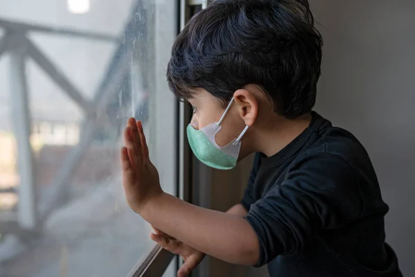 Maskeli 3 yaşında bir çocuk pencereden dışarı bakıyor.