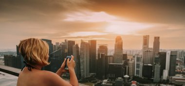 Kadın ile smartphone sonsuzluk yüzme havuzunda. İş bölgesi havadan görünümü. Marina Bay altın gün batımında Downtown manzara