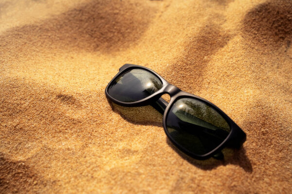 Black sunglasses on sand beach. Palm tree is reflecting on the glasses. Summer vacation concept background