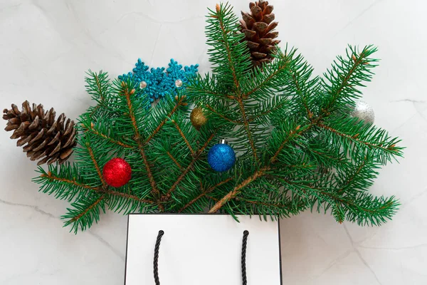 Noel 'i kutluyoruz. Noel ağacı dalları ve parlak toplardan yapılmış beyaz mermer bir arka planda beyaz bir hediye çantası..