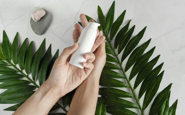Genç bir kadın beyaz bir el kremi tutuyor. Tropik yapraklar, spa konsepti. Beyaz mermer bir arka planda