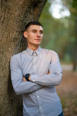 Parktaki genç bir adamın portresi, bir ağacın yanında, açık havada, doğal yeşil bir arka planda. Ciddi zeki adam uzaktan bakıp gülümsüyor. Seçici odak