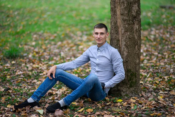 Bir sonbahar parkında, bir ağacın yanında, açık yeşil arka planda genç bir adamın portresi. Ciddi bir adam kameraya bakıp gülümsüyor. Seçici odak