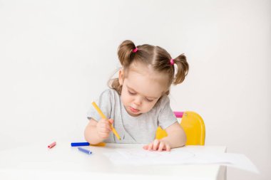 İki yaşındaki bir kızın fotoğrafı bir masada oturuyor ve beyaz arka planda renkli kalemlerle çiziyor.