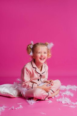 Açık pembe pijamalarla poz veren güzel bir kızın stüdyo fotoğrafı. Yastık, tüy ve saatle pembe arka planda duran neşeli bir kız bebek. Çocuk duyguları konsepti